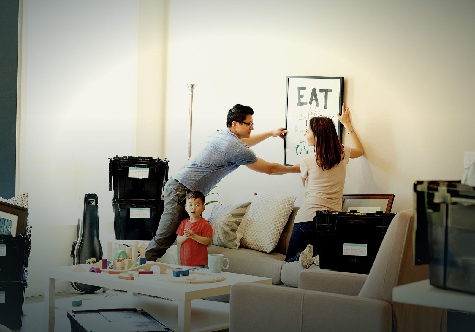 A young family hanging a picture in their new living room while their toddler plays nearby